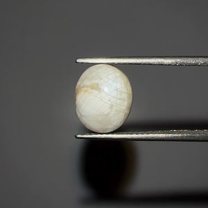 Puede incluir: Una gema ovalada, blanquecina, es sostenida por unas pinzas sobre un fondo gris oscuro. La gema tiene un brillo nacarado con patrones internos sutiles y una apariencia ligeramente translúcida. Las pinzas son de color plateado.
