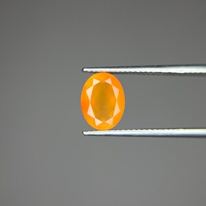 Puede incluir: Una gema naranja de forma ovalada, sujeta con unas pinzas plateadas. La gema tiene un diseño facetado con un patrón de estrella en el centro. El fondo es gris liso.