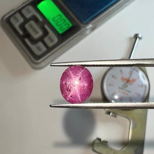 May include: A vibrant, oval-shaped ruby gemstone, held by tweezers, showcasing a star effect. A digital scale with a green display and a measuring gauge are in the background. The ruby's color is a deep pink with internal lines.