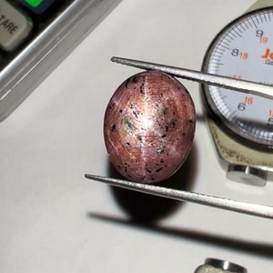 May include: A reddish-brown, oval gemstone held by tweezers, with dark inclusions and a polished surface. A gem gauge and a scale are in the background, suggesting a focus on gem measurement and evaluation.