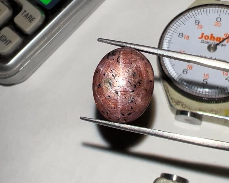 Puede incluir: Una gema ovalada de color marr&oacute;n rojizo, sujeta con pinzas, con inclusiones oscuras y una superficie pulida. Un calibrador de gemas y una balanza en el fondo sugieren la medici&oacute;n y evaluaci&oacute;n de gemas.