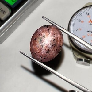 Puede incluir: Una gema ovalada, de color marr&oacute;n rosado, sujeta con pinzas. La piedra tiene motas oscuras y un brillo met&aacute;lico. Un indicador de esfera y una balanza son visibles en el fondo, lo que sugiere una medici&oacute;n.