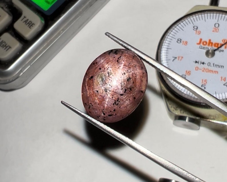 Puede incluir: Una gema ovalada, de color marr&oacute;n rosado, sujeta con pinzas. La piedra tiene motas oscuras y un brillo met&aacute;lico. Un indicador de esfera y una balanza son visibles en el fondo, lo que sugiere una medici&oacute;n.