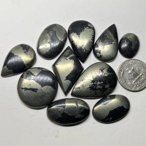 Pode incluir: Uma coleção de pedras preciosas de pirita em várias formas, incluindo gota, oval e diamante. As pedras têm uma cor cinza escuro metálico com manchas douradas. Uma moeda de um quarto de dólar dos EUA está incluída para referência de tamanho.