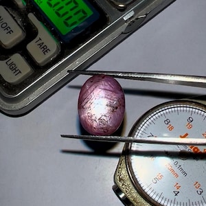 Op de afbeelding: Een roze ovaalvormige edelsteen met een stervormig patroon wordt gemeten met een schuifmaat. De schuifmaat geeft 14 mm aan. De edelsteen wordt ook gewogen op een weegschaal die 0,800 ct aangeeft.