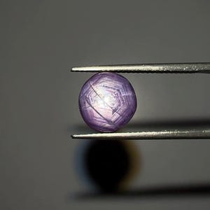 May include: A polished, oval purple gemstone held by tweezers. The gem has a star-like pattern across its surface, with light reflecting off the facets. The background is a muted grey, providing contrast to the vibrant gem.