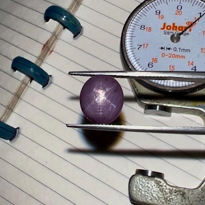 May include: A purplish-pink gemstone is measured with a gem gauge. The dial face of the gauge shows measurements. The gemstone is held between the jaws of the silver-coloured gauge, resting on lined paper. Teal clips are visible in the background.