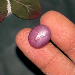 Puede incluir: Una piedra preciosa de rubí estrella pulida de forma ovalada con un tono magenta rosado intenso. La piedra tallada en cabujón muestra un efecto de estrella de seis rayos en su superficie, creado por la luz reflejada en inclusiones internas.
