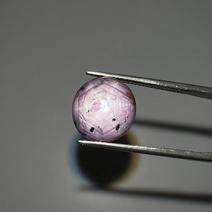 May include: A round, light purple gemstone with a star-like pattern, held by silver tweezers against a gray background. The gem has dark inclusions and a polished surface, showcasing its natural beauty. This gemstone is likely a star ruby.