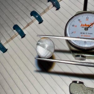 May include: A white and gray oval gemstone is held by tweezers, next to a dial gauge. The gemstone has a polished surface and is being measured. The gauge reads in millimeters, with a range of 0-20mm.