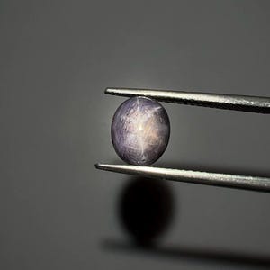 以下が含まれることがあります： ピンセットで保持された、研磨された楕円形の宝石。 宝石は紫がかったグレーの色合いで、表面には星のような効果が見られます。 ピンセットは銀色で、背景は無地のグレーです。