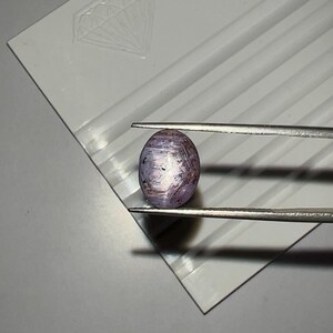 Peut inclure: Une pierre précieuse ovale, tenue par une pince, présente un mélange de teintes violettes, blanches et brunes. La gemme est posée sur une surface blanche avec un motif en forme de diamant. La surface de la pierre a un aspect texturé.