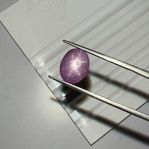 May include: A polished, oval-shaped, pink gemstone is held by tweezers against a white background. The gemstone has a star-like pattern visible on its surface. The background features a diamond-shaped design and parallel lines.