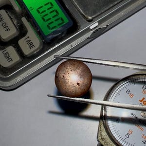 Puede incluir: Una piedra preciosa redonda e iridiscente con un esquema de color marrón y blanco se está midiendo con un calibre. El calibre lee 14 mm. La piedra preciosa también se está pesando en una báscula digital que lee 0,800 ct.