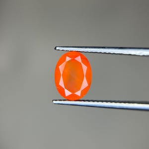 Puede incluir: Una gema facetada de forma ovalada, de un vibrante color naranja, es sostenida por unas pinzas plateadas. El tallado de la gema revela un patrón de estrella en su interior, creando un juego de luces y sombras. El fondo es gris neutro.