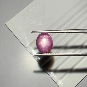 May include: A pink, oval-cut gemstone is held by tweezers against a white surface. The gem has a star-like pattern within. The tweezers are silver. The background features a diamond-shaped design. The gem is likely a ruby.