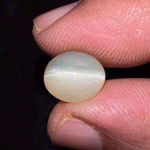 May include: A round, cabochon-cut gemstone with a translucent, off-white appearance. The stone features a distinct, horizontal chatoyant band of light across its center, creating a cat's eye effect against the smooth, polished surface of the gem.