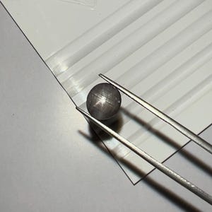 May include: A dark grey, star-shaped gemstone is held by silver tweezers against a white background with parallel grooves. The gemstone has a visible star effect, and the tweezers are positioned to showcase the stone.