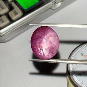 May include: A pink and white oval-shaped gemstone with a star-like pattern, held by tweezers over a measuring tool.
