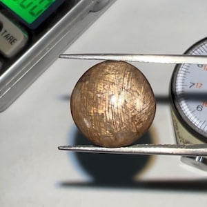 May include: A round, golden sunstone cabochon with a shimmery, metallic finish. The stone is being held by a pair of tweezers and measured with a gauge.
