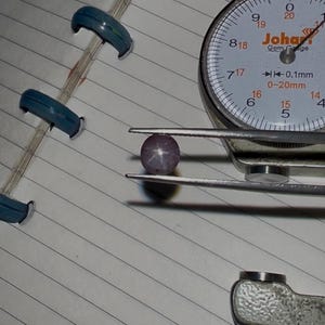 Op de afbeelding: Een close-up van een ster-robijn edelsteen die door een pincet wordt vastgehouden, naast een edelsteenmeter. De robijn heeft een lichtpaarse kleur met een stervormig patroon. De meter heeft een witte wijzerplaat met zwarte cijfers.