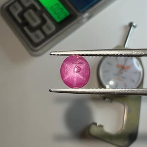 Puede incluir: Una vibrante gema rosa, de forma ovalada, es sostenida por unas pinzas. La gema se coloca frente a un indicador de esfera y una balanza digital que muestra 0,00. La superficie de la gema refleja la luz.