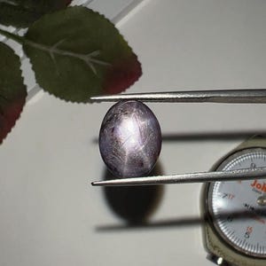May include: An oval-shaped, translucent purple star sapphire gemstone held by metal tweezers. The stone displays a distinct six-rayed star effect across its surface. A portion of a metal gem gauge is visible, showing measurements in millimeters.
