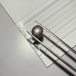 Peut inclure: Une petite pierre précieuse ronde et tachetée, tenue par une pince métallique. La gemme est de couleur gris-violet foncé avec des taches plus claires. Elle repose sur une surface blanche avec des rainures parallèles et un logo en forme de diamant.