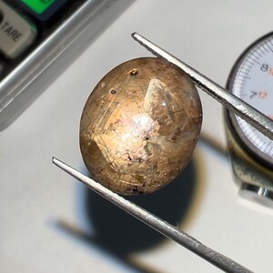 May include: A translucent, oval-shaped gemstone with a golden-brown hue is held by tweezers. The gem has internal inclusions and is being examined with a scale and a measuring gauge in the background. The gem is likely a precious stone.