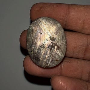 Peut inclure: Une pierre précieuse ovale polie, présentant un mélange de gris, de violet et de dorée. La pierre a une surface texturée avec des lignes et des motifs visibles. Le bijou est tenu dans une main, mettant en valeur sa forme.