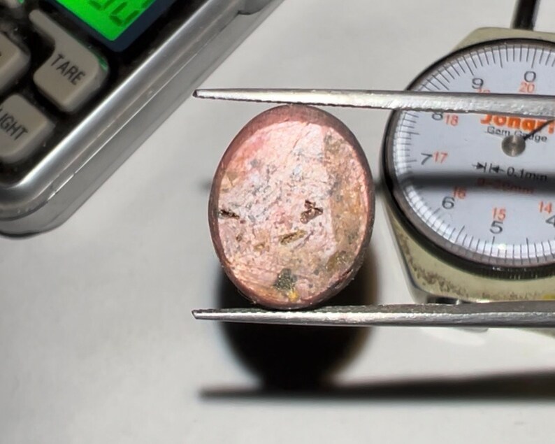 Puede incluir: Una gema en bruto de forma ovalada, sujeta con pinzas, con un tono marr&oacute;n rosado y una superficie rugosa y texturizada. Una balanza digital y un calibrador de gemas en el fondo sugieren medici&oacute;n.