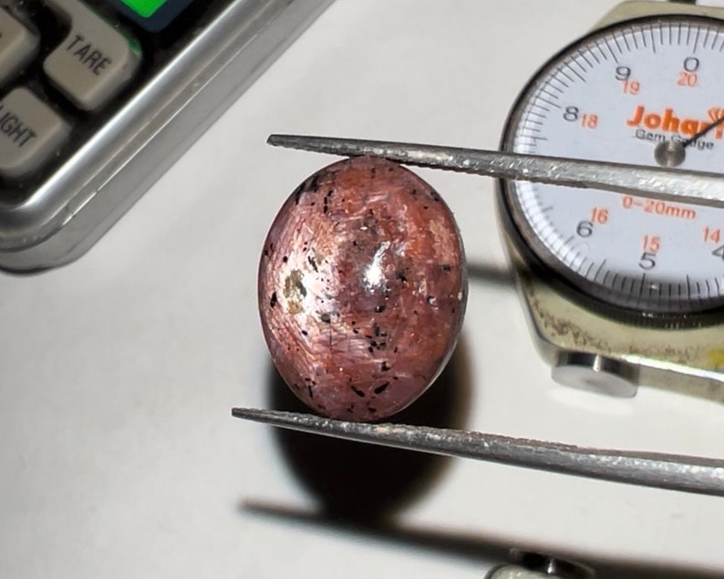 Puede incluir: Una gema ovalada pulida, que muestra inclusiones rosas, marrones y negras. La gema est&aacute; sujeta con pinzas, con un calibrador de gemas y una balanza en el fondo. La superficie de la gema tiene una apariencia texturizada.