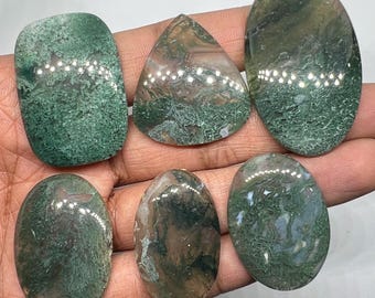 May include: A collection of six polished moss agate gemstones in various shapes, including oval, rectangular, and triangular. The stones display a mix of green, brown, and white patterns, with a natural, earthy appearance. The gemstones are held in a hand.