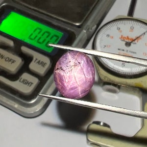 May include: A close-up of a pink and purple gemstone being measured with a caliper. The gemstone is round and has a striated surface. The caliper reads 7.5 mm.