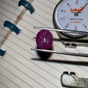 May include: A polished, deep pink gemstone is held by a metal caliper. The caliper is positioned over a lined notebook page. A dial gauge with the brand name "Johari" is visible, indicating measurements in millimetres.