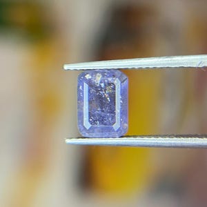 May include: A rectangular, faceted gemstone held by tweezers. The gem is a deep blue colour with iridescent inclusions. The cut is an emerald shape, with a rectangular face and bevelled edges. The background is blurred, with yellow and brown tones.