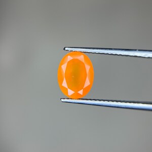 Puede incluir: Una gema naranja de forma ovalada sostenida por unas pinzas sobre un fondo gris. La gema tiene un diseño facetado, con cortes triangulares que irradian desde el centro. Las pinzas son de color plateado.