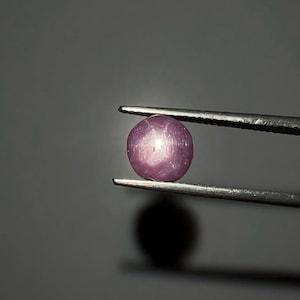 May include: A round, polished, purplish-pink gemstone is held by a pair of tweezers against a dark gray background. The gem has a smooth surface and a subtle, star-like effect. The tweezers are silver-colored.