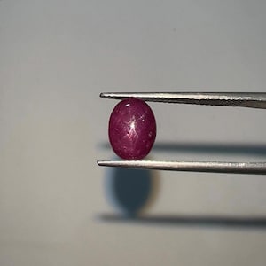 Puede incluir: Una gema de color rojo intenso, de forma ovalada, es sostenida por unas pinzas plateadas. La gema tiene una superficie pulida y parece translúcida. El fondo es de color gris claro, proporcionando un telón de fondo neutro para la gema.