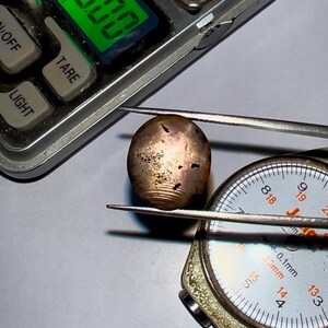 May include: A close-up of a gemstone being measured with a caliper. The gemstone is a light brown color with a shimmery finish. The caliper reads 14.4 mm.