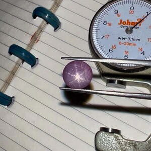 May include: A purplish-pink gemstone is held by a gem gauge. The gem gauge has a white dial with black numbers and the text "Johar Gem Gauge". The gemstone has a star-like pattern on its surface. The gauge measures in mm.