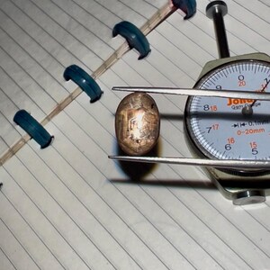 Puede incluir: Una gema ovalada de color cobre se mide con un indicador de cuadrante. El indicador muestra milímetros y la gema se sujeta con pinzas. Un cuaderno con anillas azules está en el fondo.