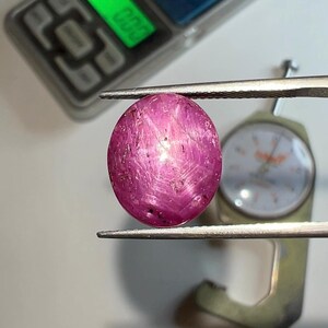 Puede incluir: Una gema de rubí ovalada y pulida sujeta con pinzas. La gema es de color rosa intenso con estrías internas. Una balanza digital con pantalla verde y un medidor se encuentran en el fondo.
