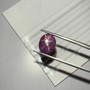 May include: A polished, oval-shaped, purplish-red gemstone is held by tweezers against a white background. The gem displays a star-like effect. The background features a diamond-shaped design and parallel lines. This image showcases a precious stone.