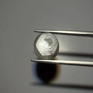 May include: A round, faceted gemstone held by tweezers. The gem is a pale, translucent gray color with internal fractures. The tweezers are silver-colored and positioned against a gray background, highlighting the gem's shape and detail.