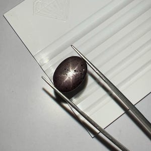 Könnte beinhalten: Ein dunkler, ovaler Edelstein mit sternförmigem Muster, gehalten von silbernen Pinzetten. Der Edelstein hat eine tiefviolette Farbe und liegt auf einer weißen Oberfläche mit einem rautenförmigen Logo. Die Pinzetten sind angewinkelt.