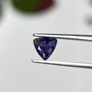 May include: A triangular gemstone held by tweezers. The gem is a deep indigo colour with internal flecks of lighter blue and purple. The shape is an equilateral triangle, showcasing the gem's polished surfaces and clarity.