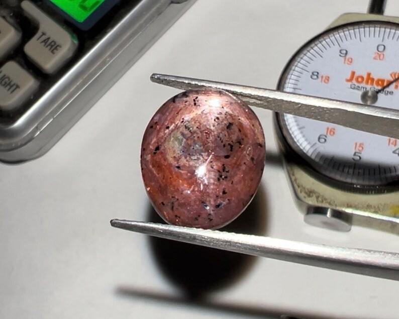 Puede incluir: Una gema pulida de forma ovalada sujeta con pinzas. La gema es una mezcla de marr&oacute;n rojizo y rosa con inclusiones oscuras. Un calibrador est&aacute; en el fondo. La gema se est&aacute; midiendo.