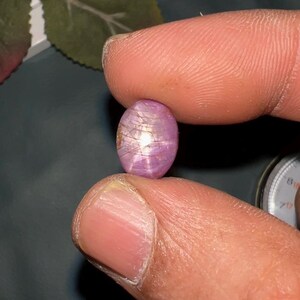 May include: An oval-shaped, translucent pinkish-purple star sapphire gemstone featuring a distinct six-rayed star pattern across its surface. The cabochon-cut stone displays internal inclusions and a bright light reflection at its center point.