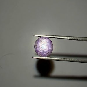 May include: A spherical, lilac-hued gemstone held by tweezers. The gem displays a star-like effect across its surface. The background is a neutral grey, accentuating the gem's colour and form. This image presents a precious stone, possibly for jewellery.